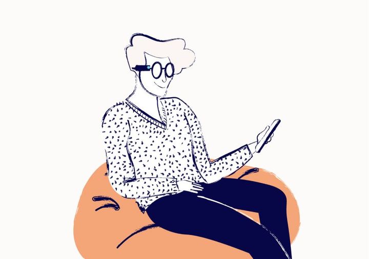 A person in a spotty sweater sits on a bean bag. They are wearing assistive tech on their glasses and are looking at a phone