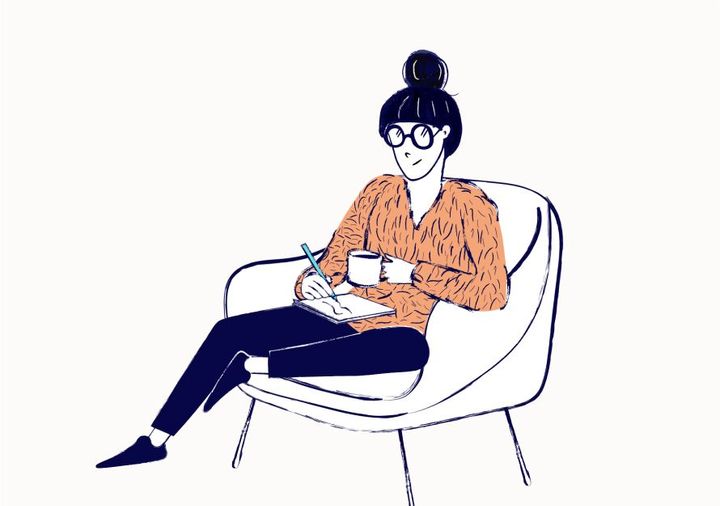 A young woman in glasses sits on an armchair. She is writing in a notebook and holding a mug of coffee.